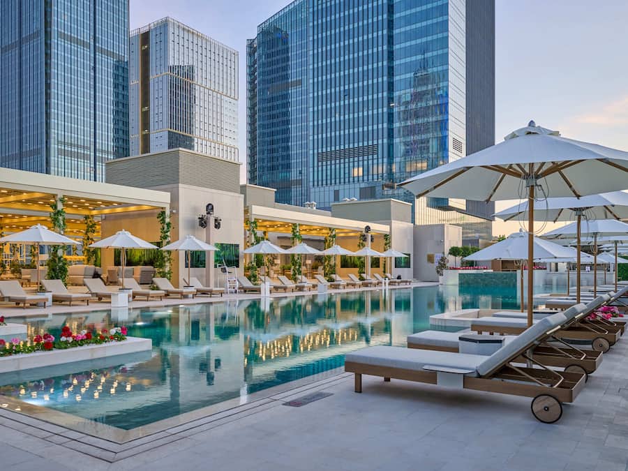 Una piscina en la azotea con sillas de descanso y sombrillas, rodeada de modernos edificios altos.
