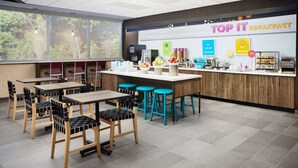 A modern breakfast area with wooden tables and chairs, a counter with various food items, and a large window with a view of trees. - Tru by Hilton Atlanta Northlake Parkway, GA (Atlanta)