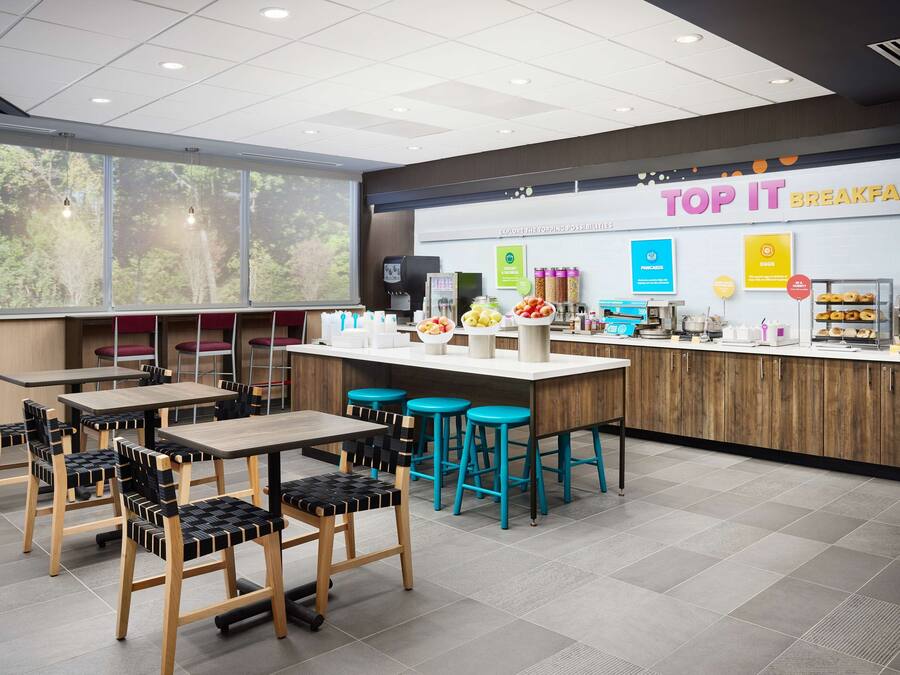 A modern breakfast area with wooden tables and chairs, a counter with various food items, and a large window with a view of trees.