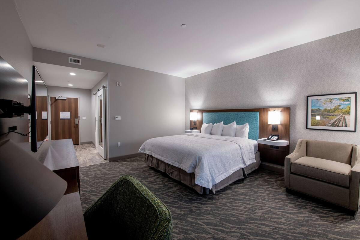 A hotel room with a bed, a chair, a desk, and a painting on the wall.
