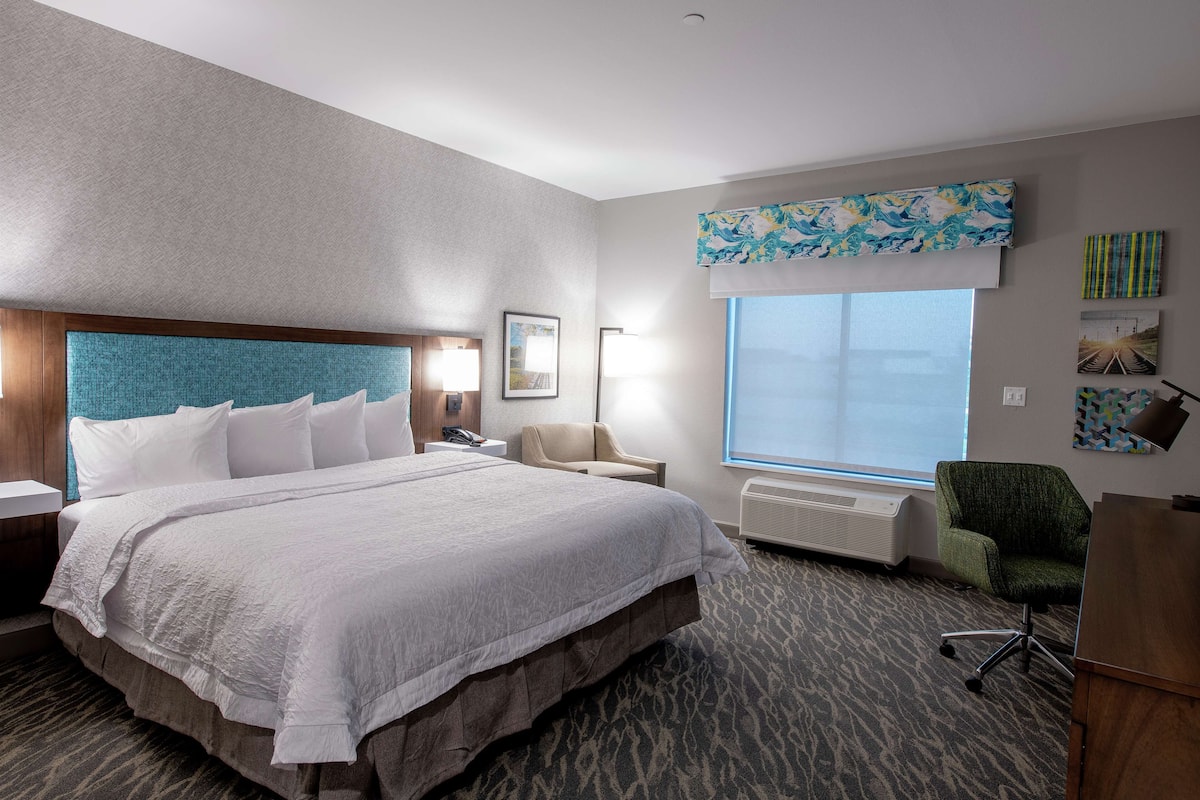 A hotel room with a large bed, a desk, a chair, a window with a view, and a wall with artwork.