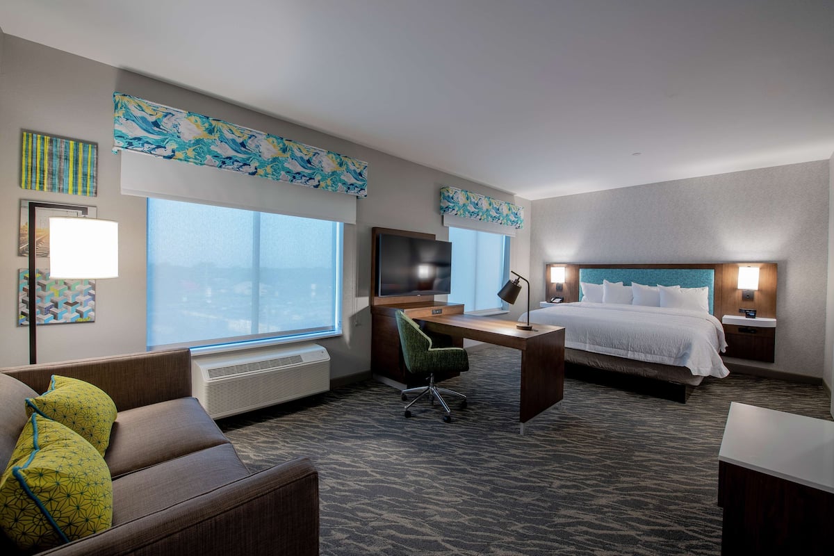 A hotel room with a bed, desk, chair, and large windows.