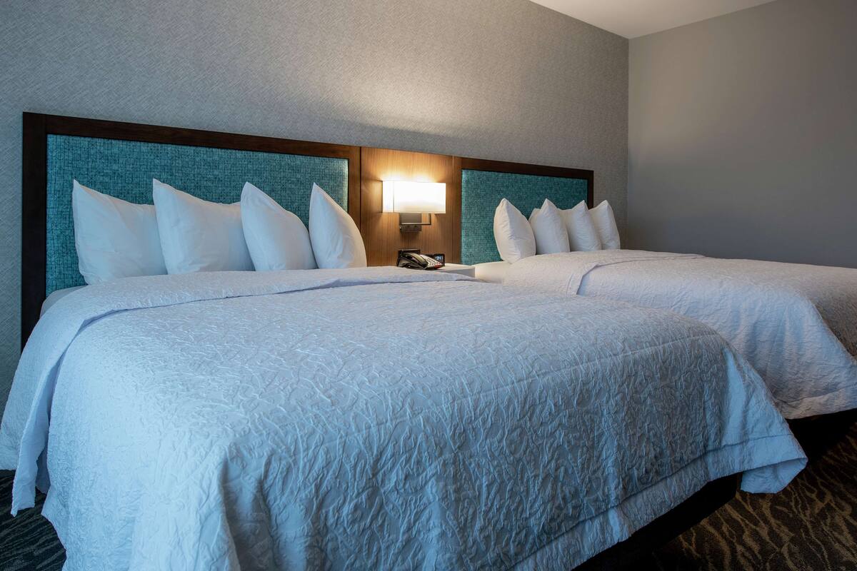 A hotel room with two beds, a headboard with a teal panel, and a bedside table with a lamp.
