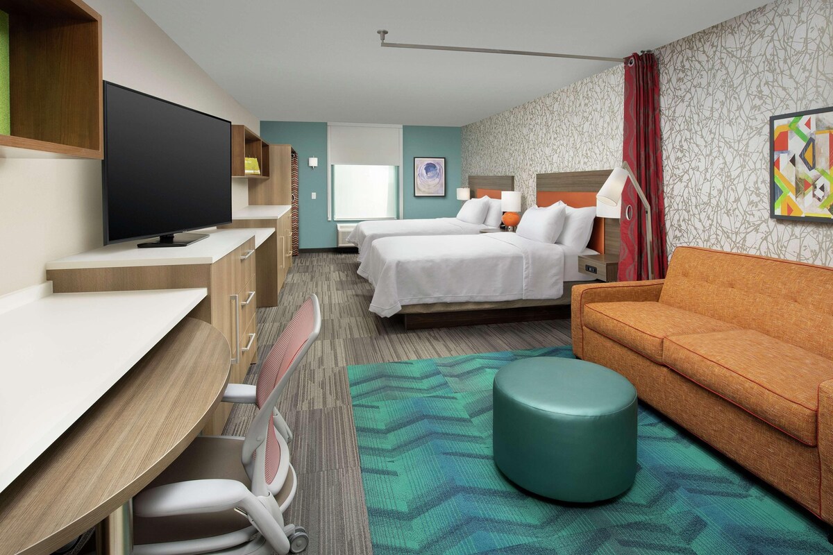 A hotel room with a bed, a desk with a television, a sofa, and a small ottoman.