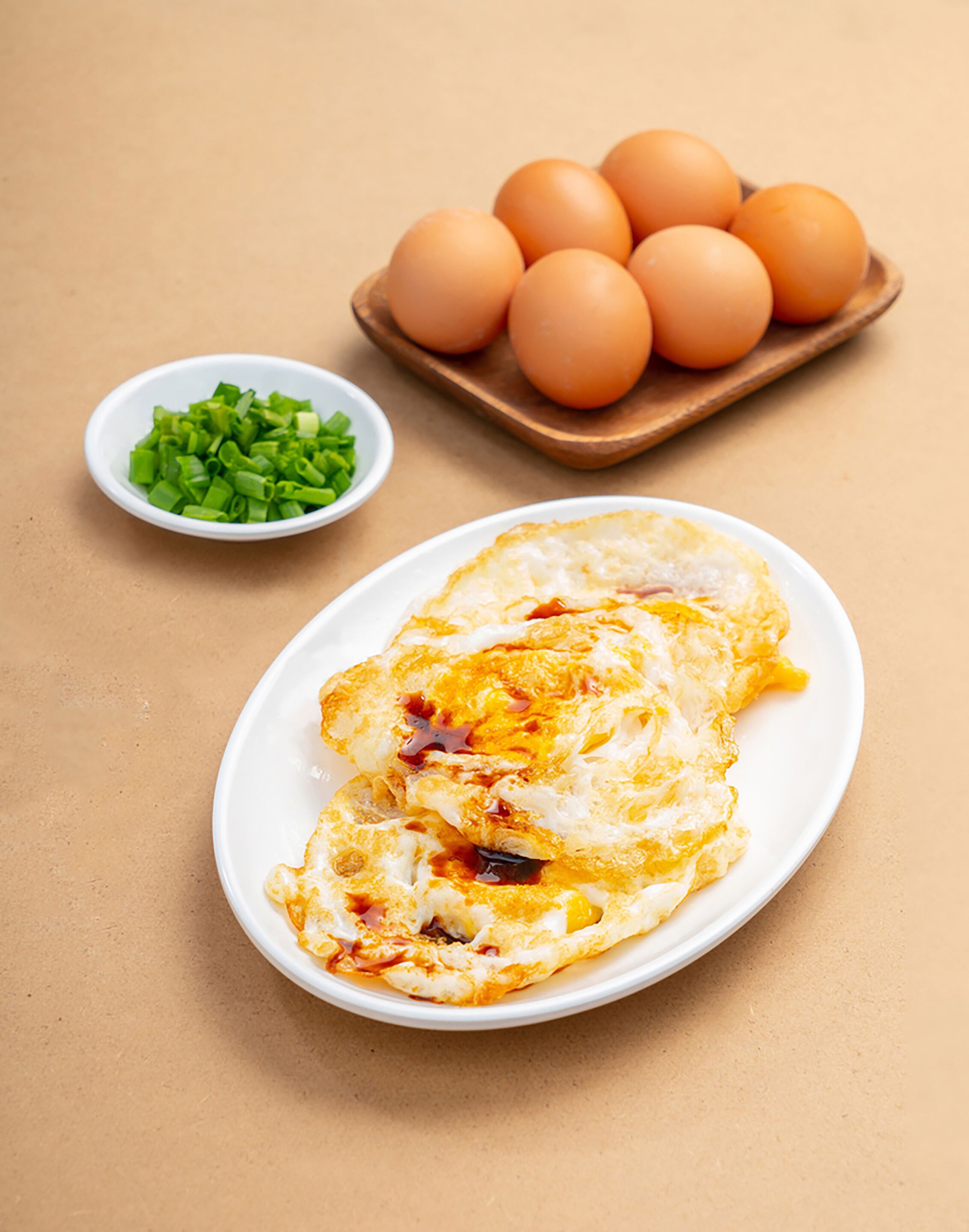 A plate with two sunny-side-up eggs, a bowl of chopped green onions, and a wooden tray with six eggs.