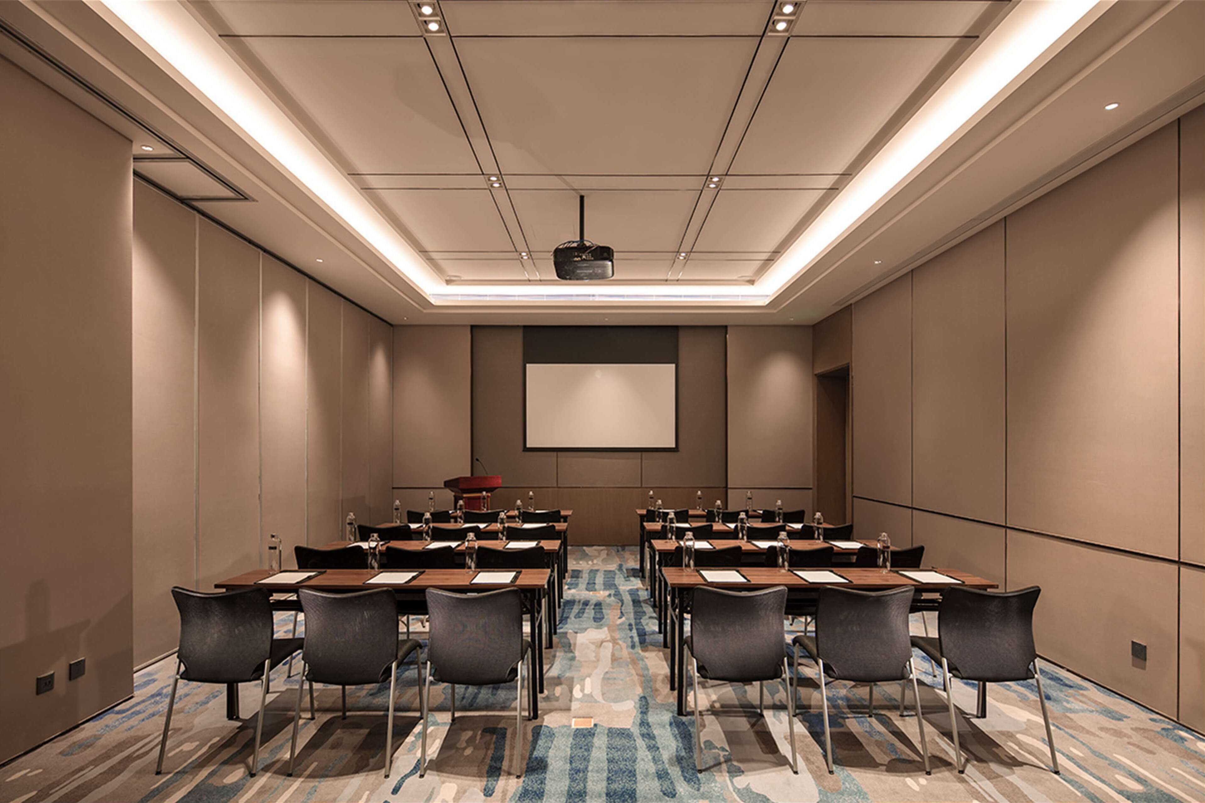 A conference room with a large screen, rows of tables and chairs, and a carpeted floor.