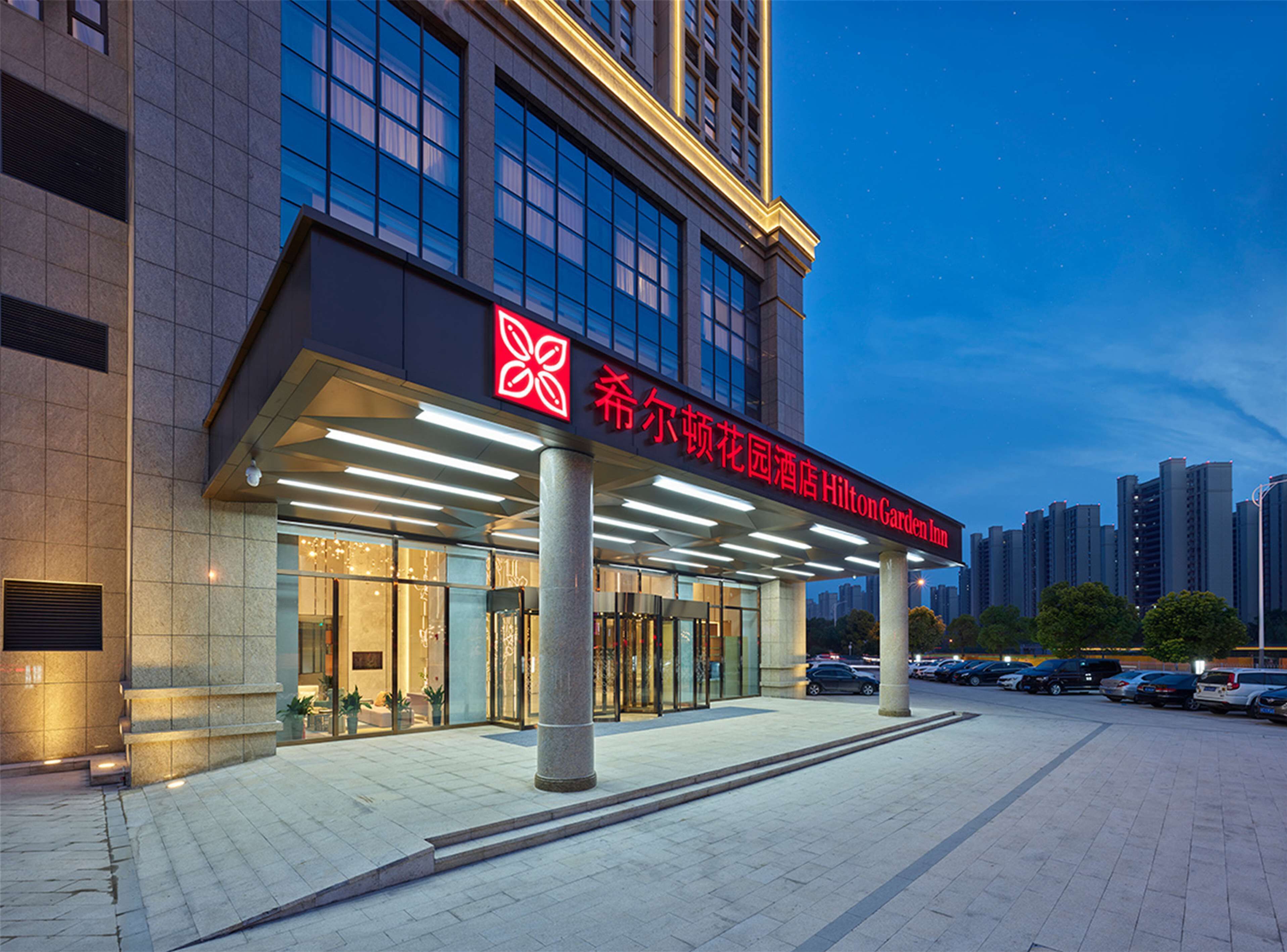 The exterior of the Hilton Garden Inn with a prominent red sign, large windows, and a parking area.