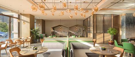 A modern restaurant interior with round tables, cushioned seating, hanging pendant lights, and a visible kitchen area in the background.