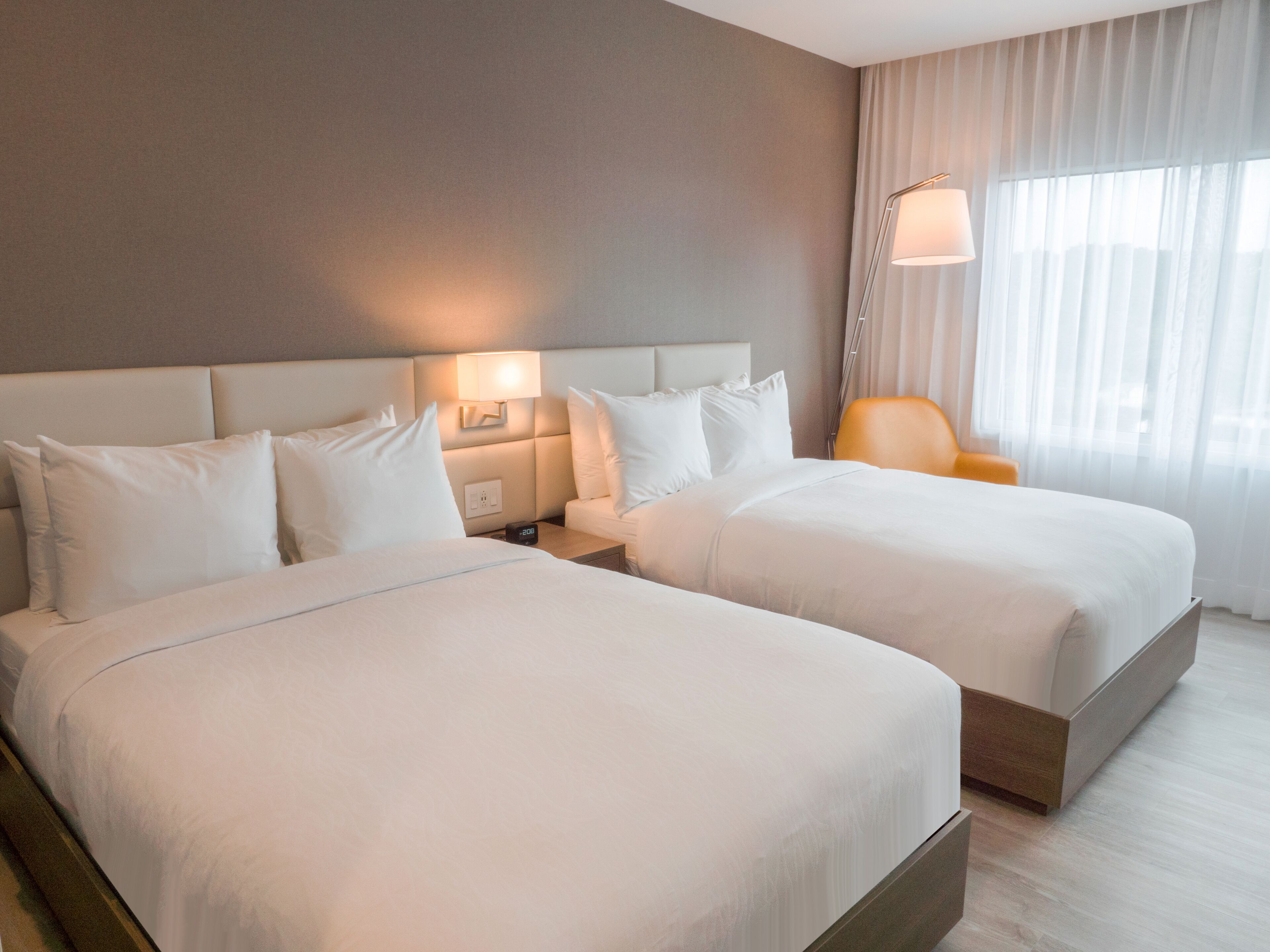 A hotel room with two beds, a nightstand with a lamp, a chair, and a large window with curtains.