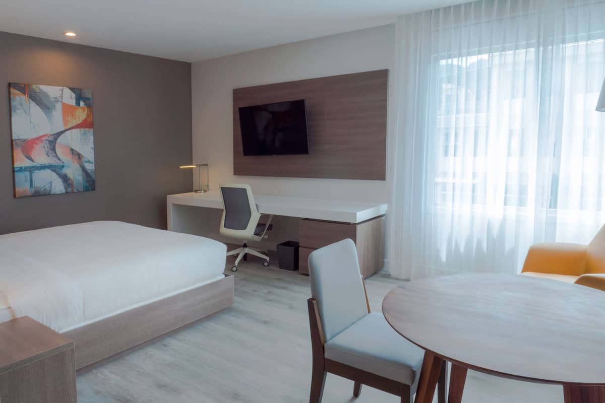 Una habitación de hotel con una cama, un escritorio con silla, un televisor montado en la pared y una mesa de comedor con sillas.