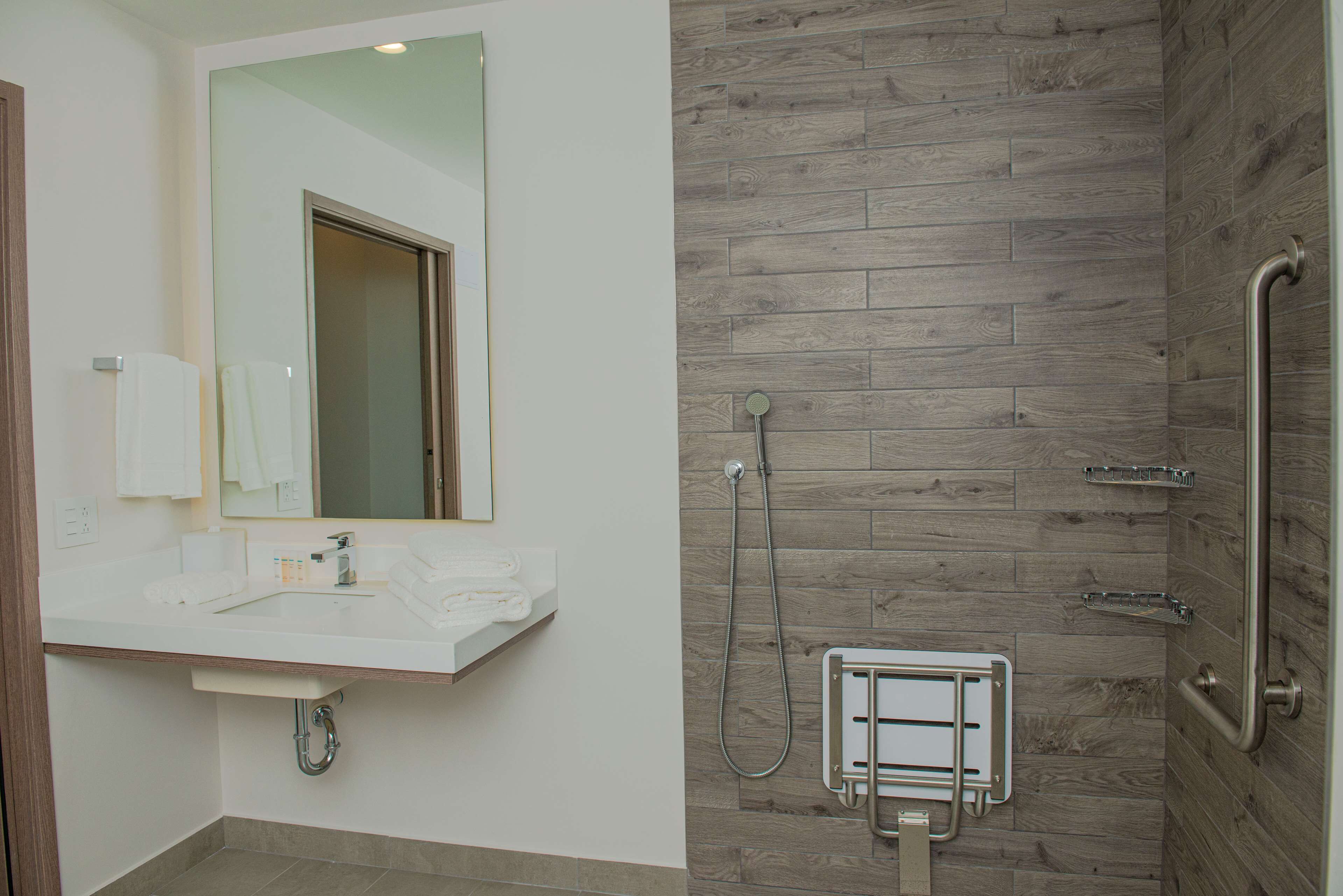 Un baño moderno con pared de ducha de madera, un lavabo blanco con espejo y una barra de apoyo en la ducha.