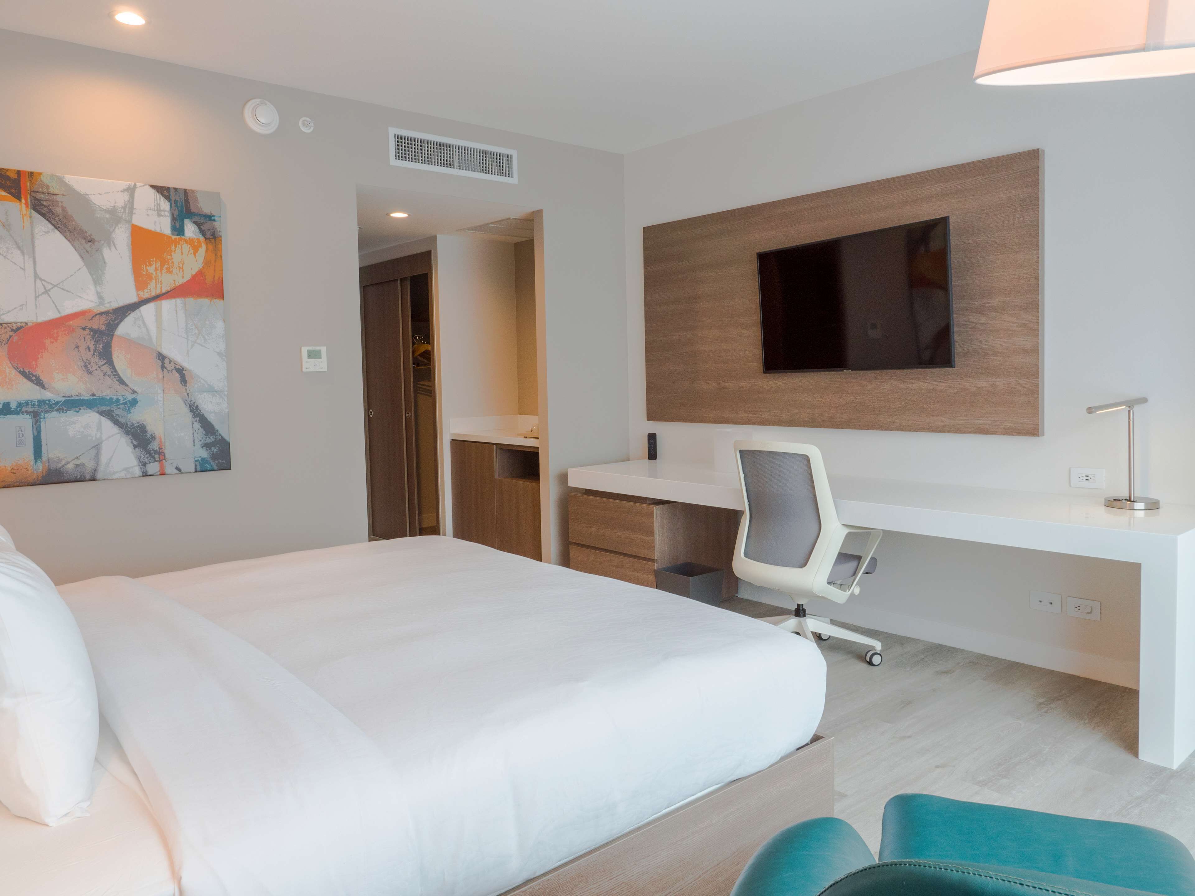 A modern hotel room with a large bed, a desk with a chair, a flat-screen TV mounted on the wall, and a painting on the wall.