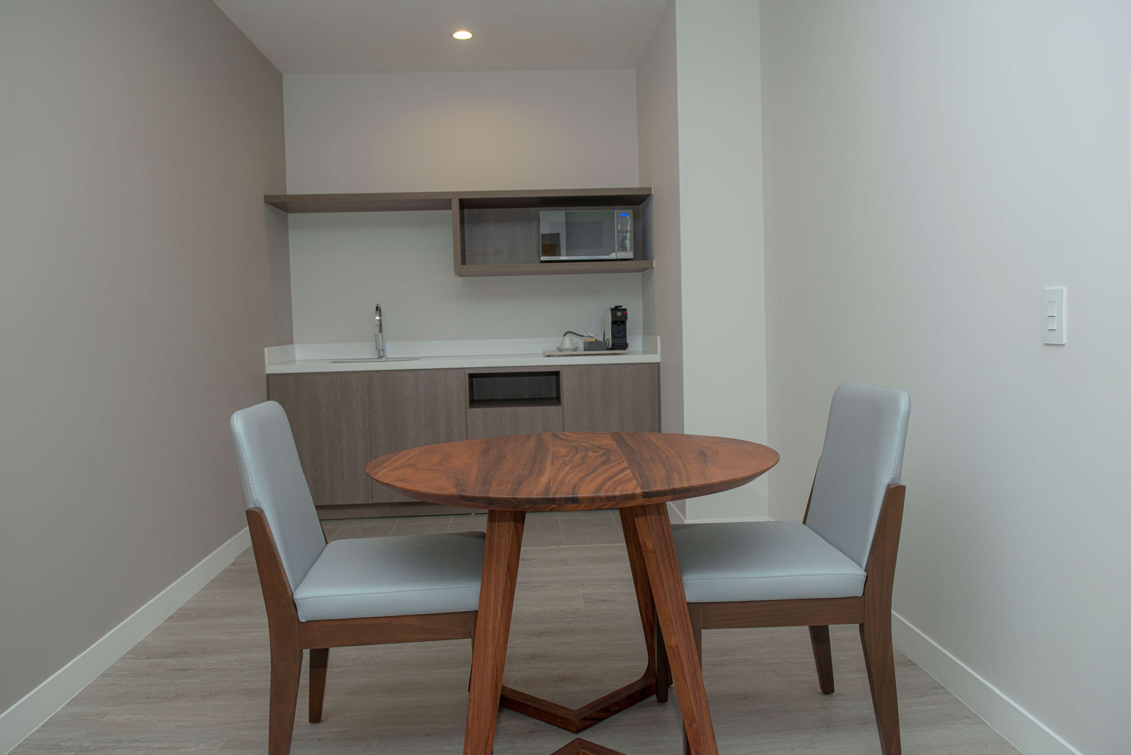 A small kitchen with a microwave, a coffee maker, and a sink. There is a round wooden table with two chairs in front of the kitchen.