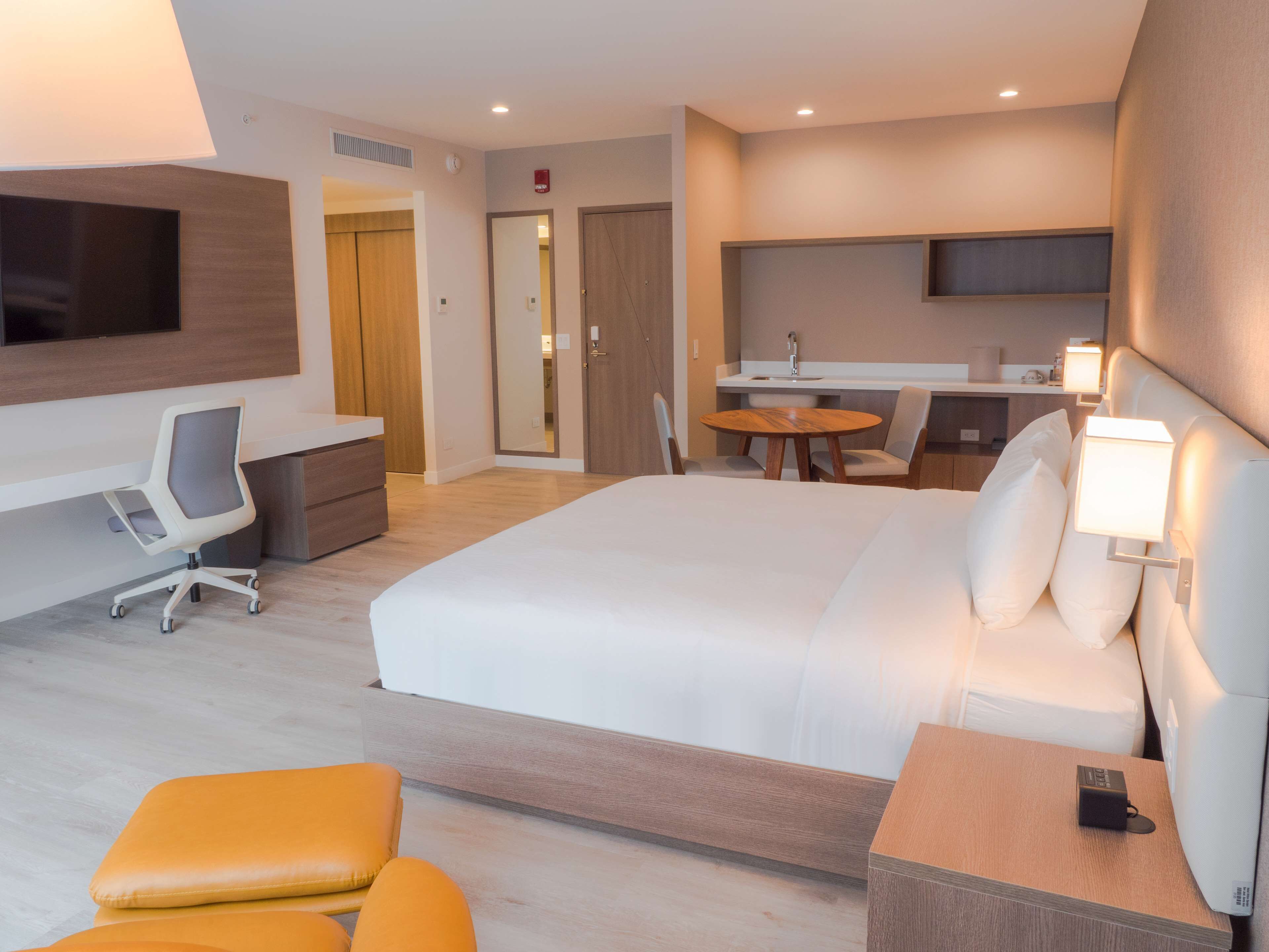 Una habitación de hotel moderna con una cama grande, un escritorio con una silla, una mesita redonda con sillas, un televisor montado en la pared y una mesita de noche con una lámpara.