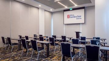 A conference room with tables and chairs, a projector screen displaying Hilton Garden Inn Santa Ana logo, and a patterned carpet.