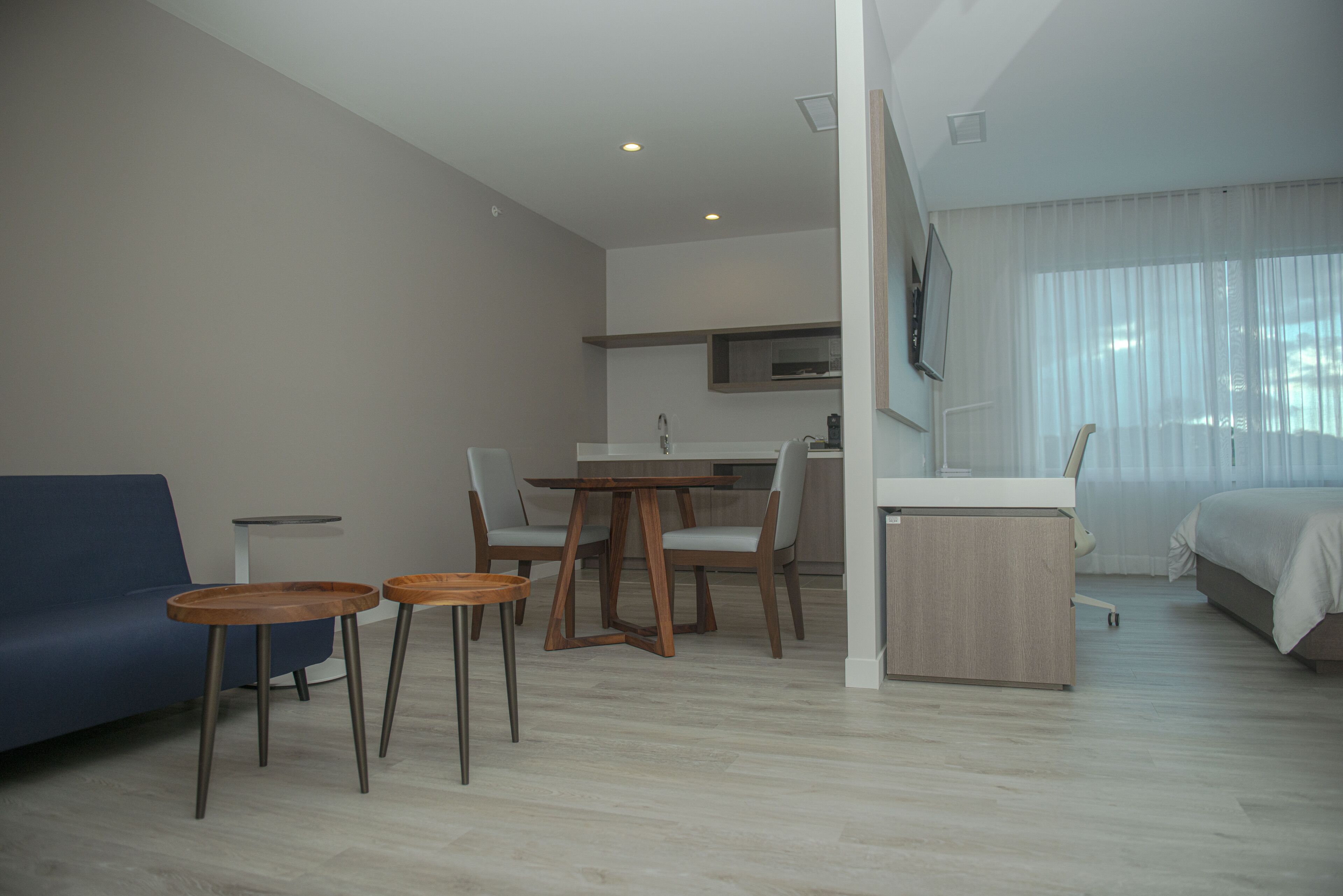 Una habitación de hotel moderna con sofá, una pequeña mesa de comedor con sillas, kitchenette, cama y un ventanal con cortinas translúcidas.