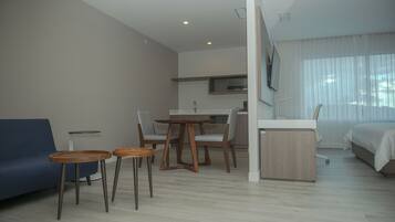 Una habitación de hotel moderna con sofá, una pequeña mesa de comedor con sillas, kitchenette, cama y un ventanal con cortinas translúcidas.