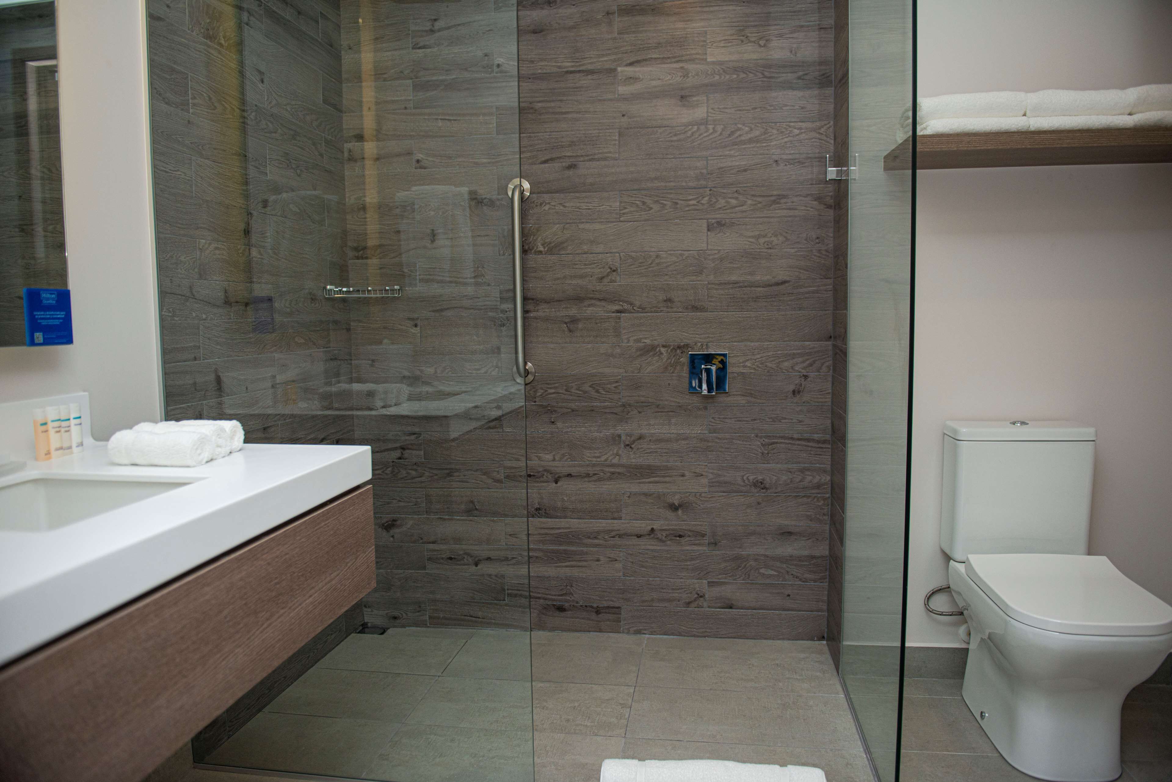 Un baño moderno con cabina de ducha de vidrio, panel de pared de madera, inodoro blanco y tocador con espejo y barra para toallas.