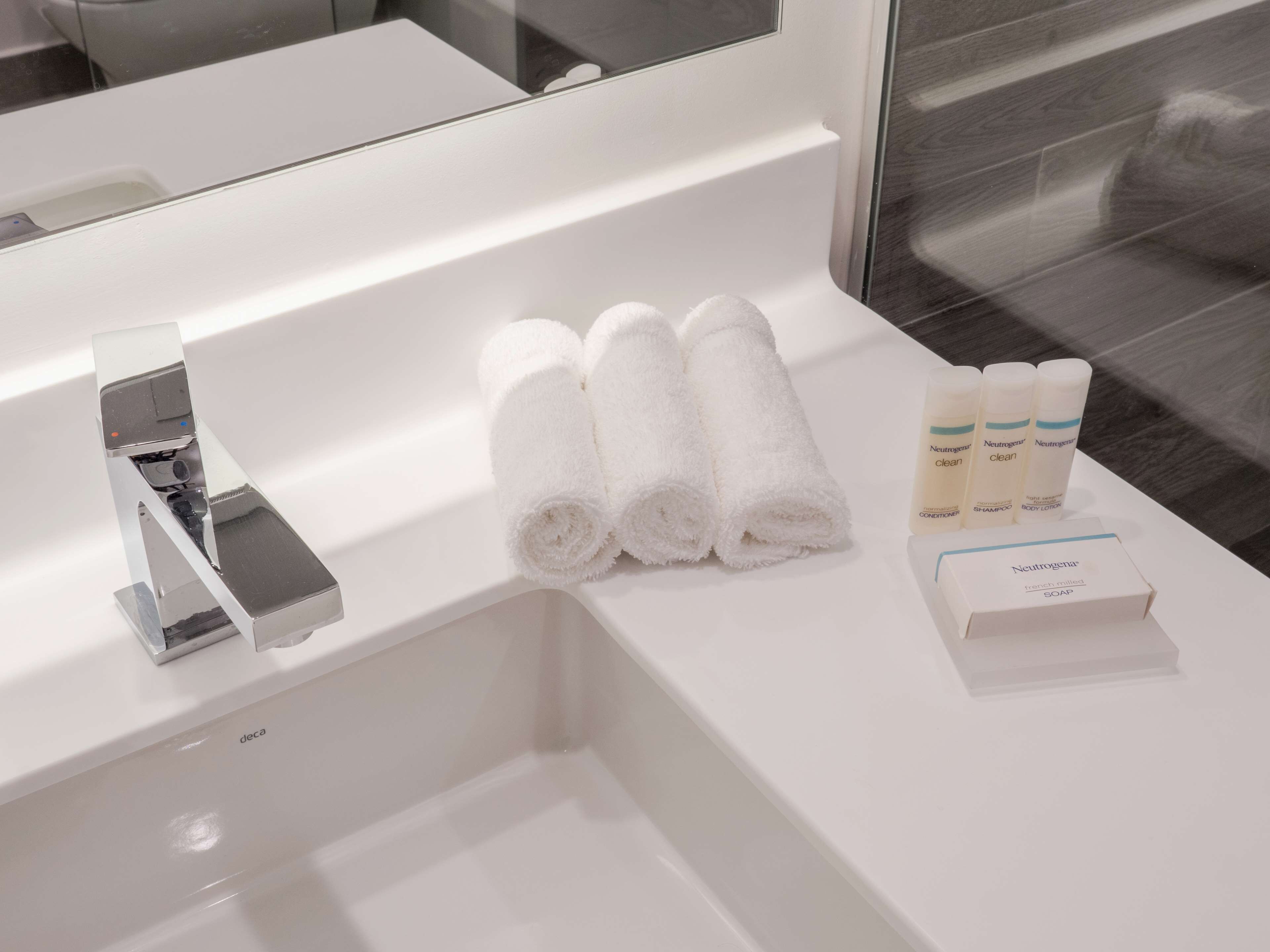 A bathroom with a white sink, a chrome faucet, and a mirror above it. There are three rolled white towels and three bottles of Neutrogena products on the sink.