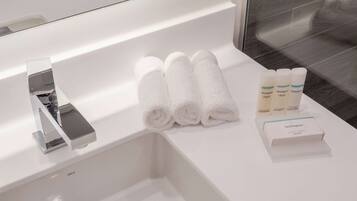 A bathroom with a white sink, a chrome faucet, and a mirror above it. There are three rolled white towels and three bottles of Neutrogena products on the sink.