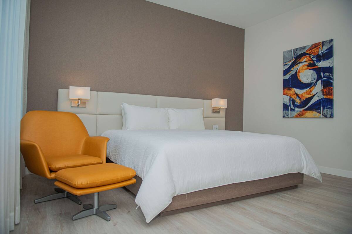 Una habitación de hotel moderna con una cama grande, un sillón naranja brillante y un cuadro en la pared.
