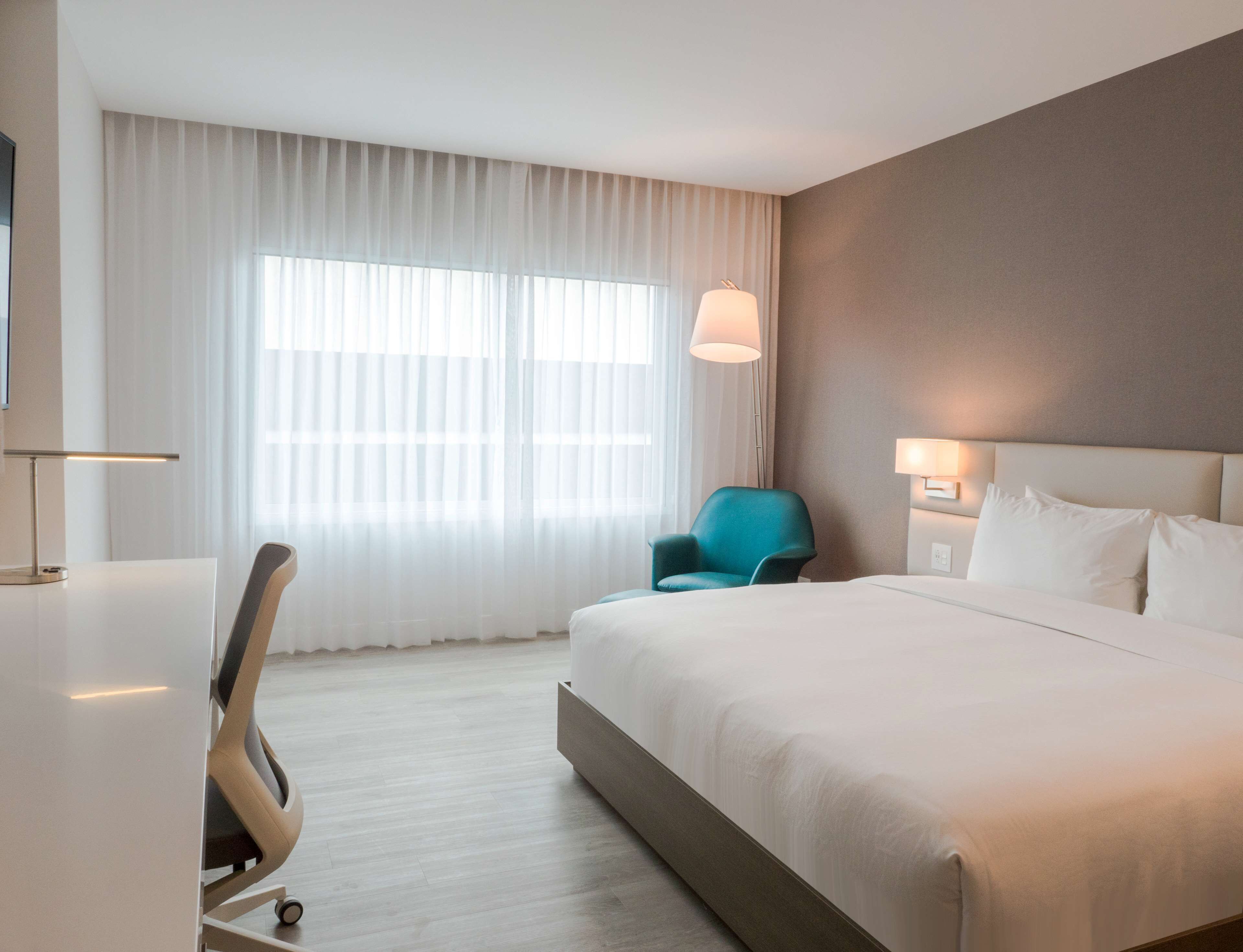 Une chambre d’hôtel avec un grand lit, un bureau avec une chaise, une fenêtre avec des rideaux légers et une lampe fixée au mur.