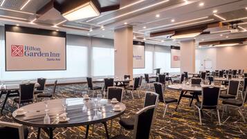 Un salón de conferencias en Hilton Garden Inn Santa Ana con varias mesas y sillas, ventanas amplias y un diseño moderno en el techo.