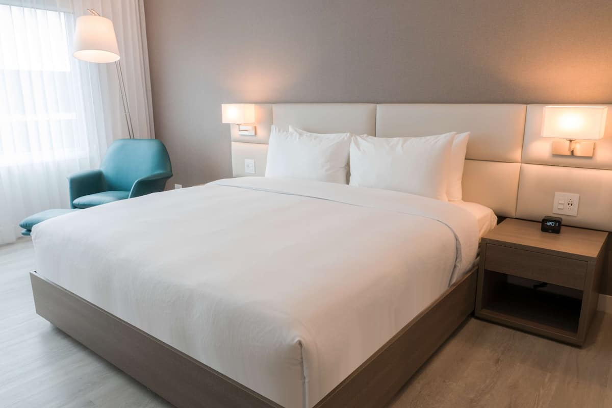 Una habitación de hotel moderna con una cama grande, una mesita de noche con reloj digital, una silla y una lámpara.