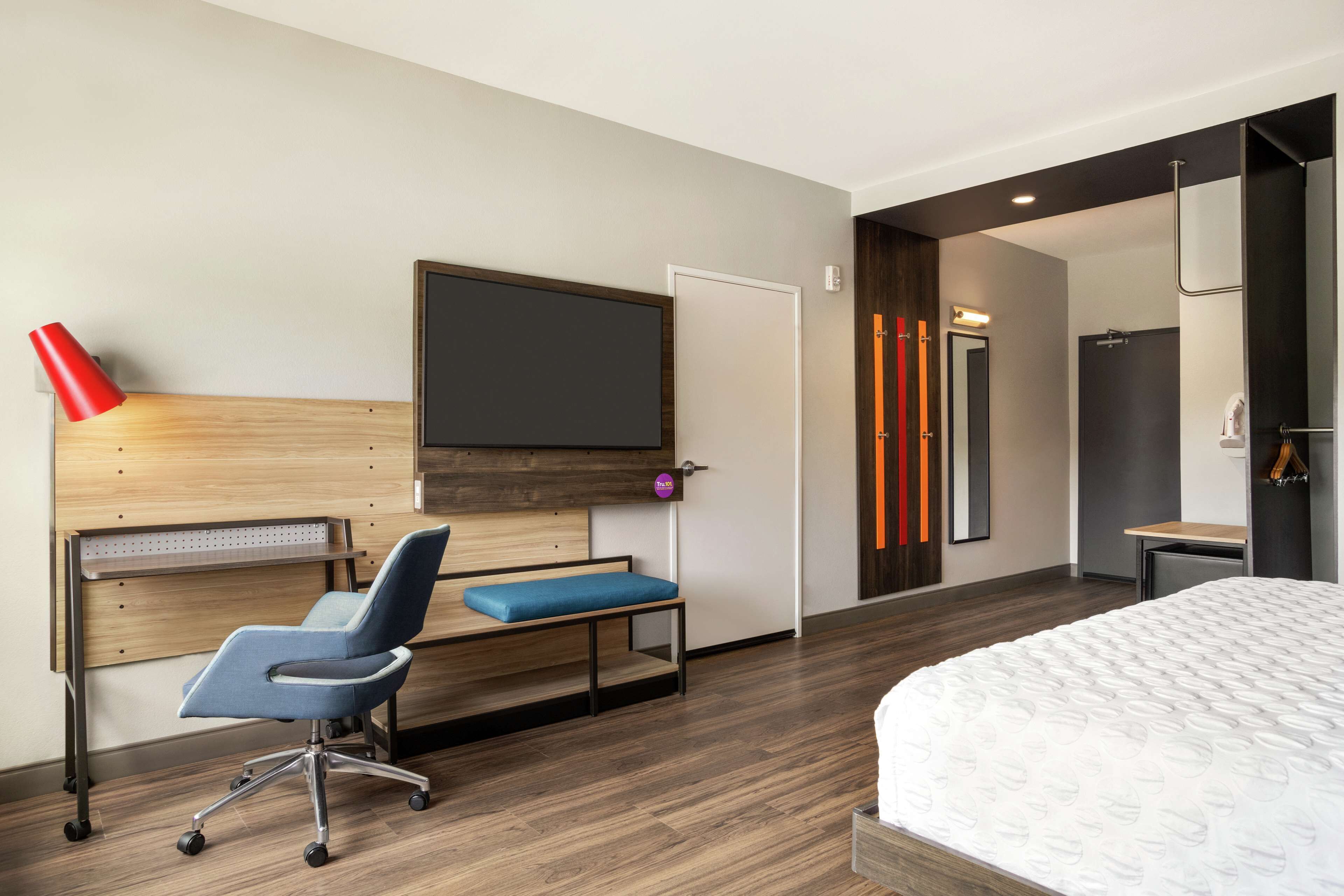 A modern hotel room with a wooden headboard, a flat-screen TV, a blue office chair, a bed with a white mattress, and a desk with a red lamp.