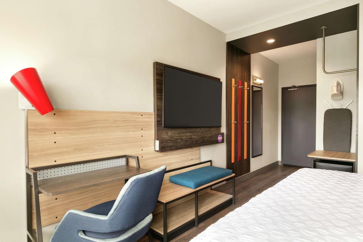 Ett modernt hotellrum med en sänggavel i trä, en platt-TV, ett skrivbord med en stol, en säng med en vit madrass och en duschhörna.
