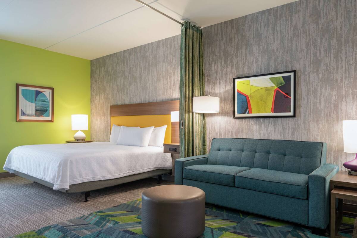Una habitación de hotel con cama, sofá, otomana redonda y un cuadro en la pared.