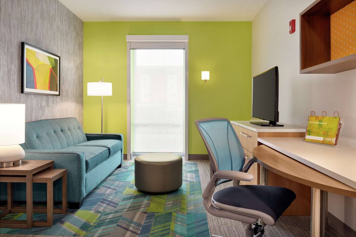 Habitación de hotel con sofá azul, un otoman redondo, un escritorio con silla, televisor y lámpara.