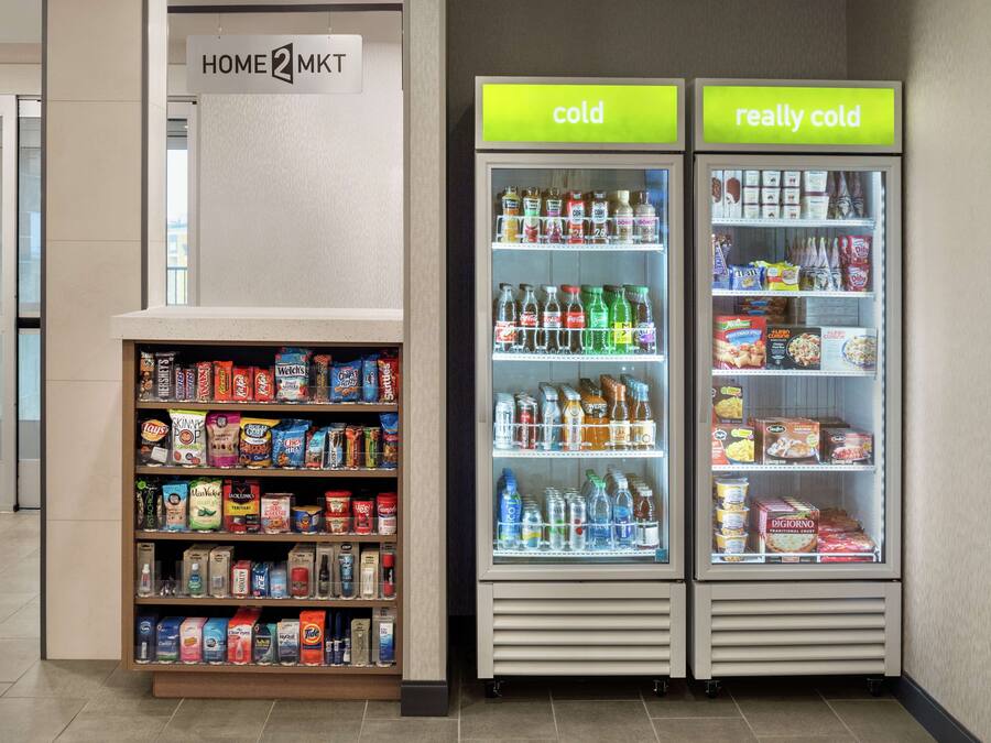 Uma área com máquinas de venda automática, com um letreiro 'HOME 2 MKT' acima e dois refrigeradores de exibição com as etiquetas 'frio' e 'muito frio'.