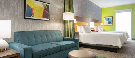 Una habitación de hotel con un sofá azul, una cama con ropa blanca, una cortina verde y una alfombra colorida.