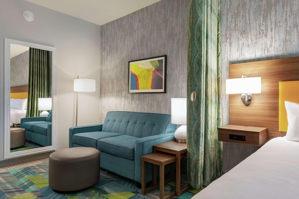 Une chambre d’hôtel avec un canapé de couleur turquoise, un petit pouf, une table de chevet en bois avec une lampe, un lit avec une tête de lit en bois et un miroir qui reflète la pièce.