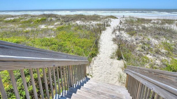 Eine Holztreppe führt zu einem Sandstrand mit vereinzelter Vegetation.
