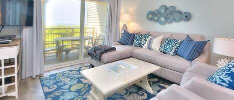 A living room with a sofa, coffee table, and a TV. There is a balcony with outdoor furniture visible through the glass doors.