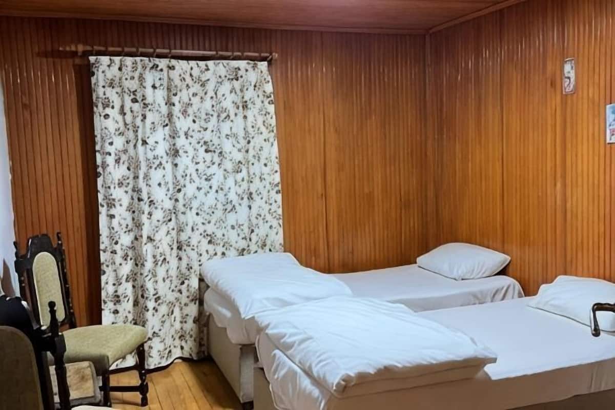A room with wooden paneling, two beds with white linens, a floral curtain, and a chair.
