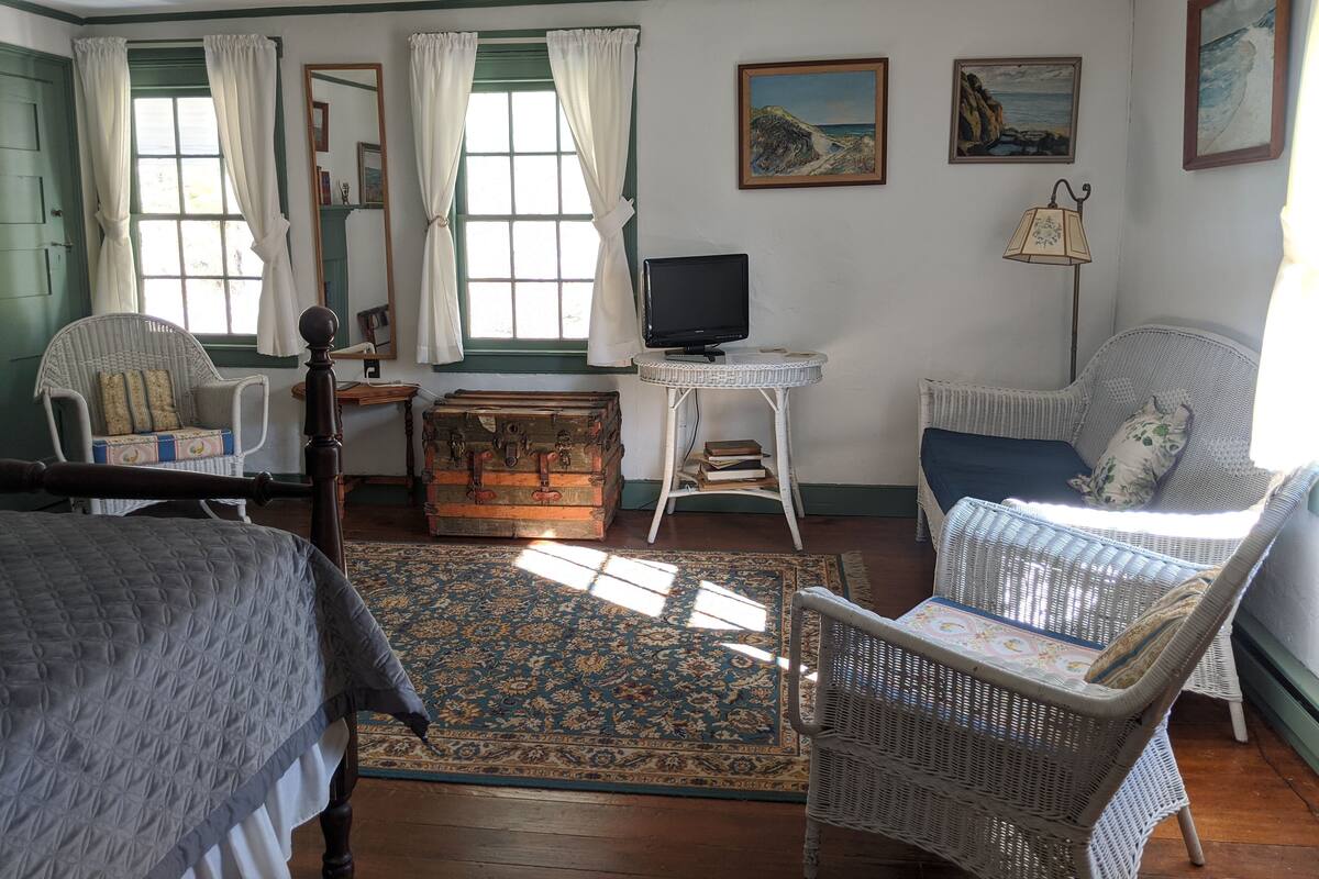 A cozy room with a bed, two wicker chairs, a small table with a television, a wooden chest, and a patterned rug.