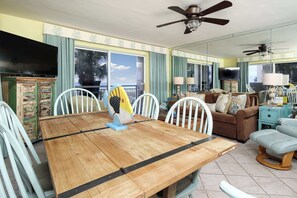 A dining area with a wooden table, white chairs, and a fish-shaped centerpiece. There is a ceiling fan, a TV, and a sofa in the background. - Pelican Isle 101🌊Gulf Front🏖️Free Beach Setup (Fort Walton Beach)