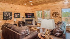 A wooden cabin interior with a fireplace, leather sofas, a television, and framed artwork on the walls.