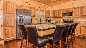 A kitchen with wooden cabinets, a stainless steel refrigerator, a microwave, a granite countertop, and four leather bar stools.