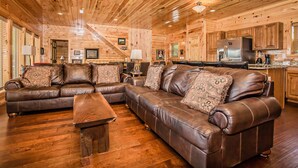A wooden interior with a brown leather sectional sofa, a wooden coffee table, and a kitchen area with wooden cabinets and appliances.