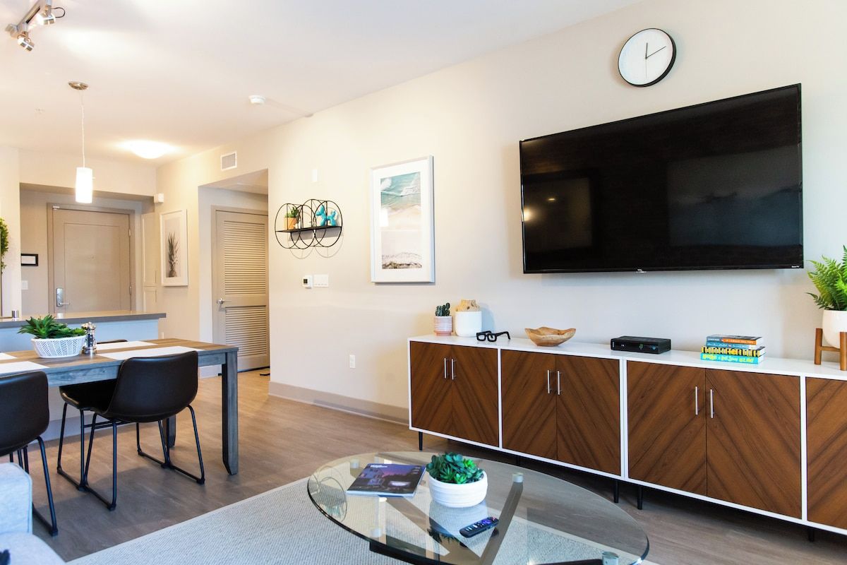 A modern living room with a flat-screen TV, a dining area with a table and chairs, and a kitchen in the background.