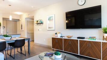 A modern living room with a flat-screen TV, a dining area with a table and chairs, and a kitchen in the background.