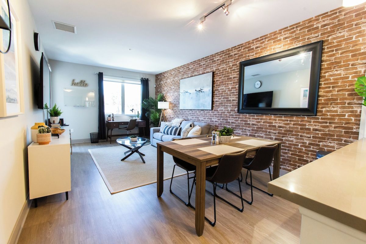 A modern living room with a brick wall, a dining area with a wooden table and chairs, and a sofa with cushions.