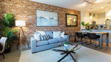 A modern living room with a brick wall, a grey sofa, a glass coffee table, and a dining area with chairs.