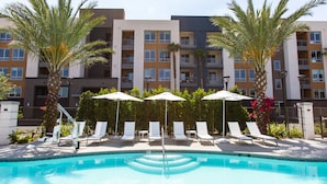 A pool area with lounge chairs and umbrellas, surrounded by palm trees and a multi-story building in the background.