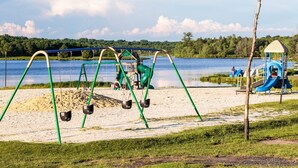 Un parque infantil con columpios y toboganes, un pequeño refugio y una zona de arena cerca de un lago.