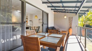 Un espace de restauration extérieur moderne, avec des tables à plateaux de verre et des chaises en bois, une cuisine en arrière-plan et un balcon offrant une vue sur la verdure.